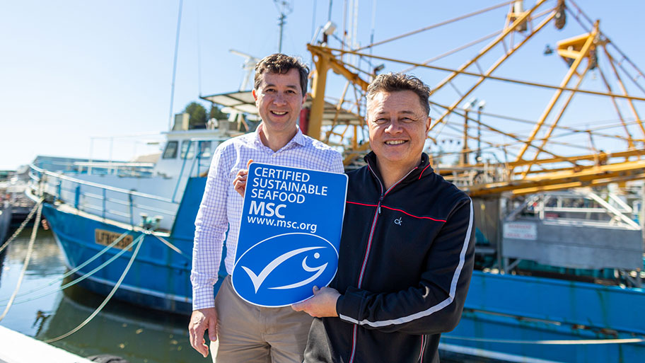 Two men in front of fishing vessel holding a sign which says: certified sustainable seafood MSC www.msc.org