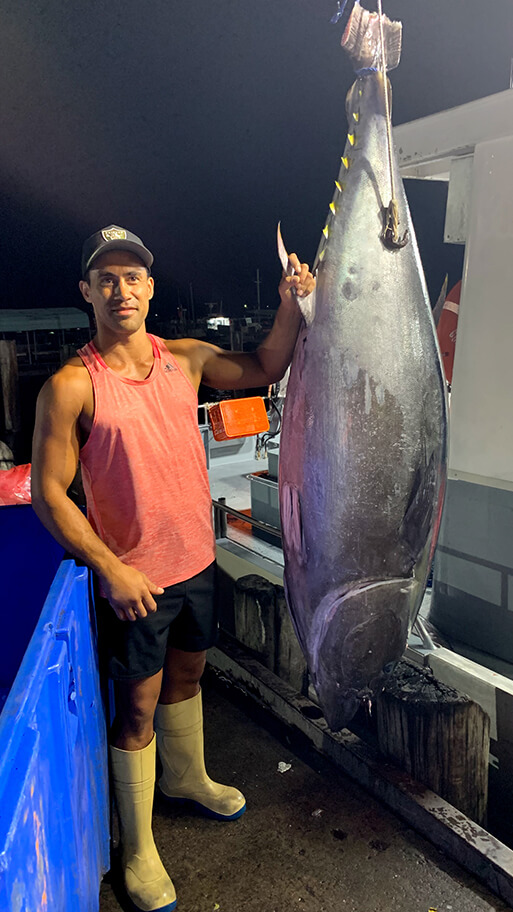A man stands next to a large tuna fish hanging from a rope as the fish is unloaded from a fishing vessel