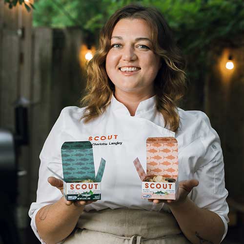 Woman wearing white chef's jacket holding two boxes of Scout brand seafood products