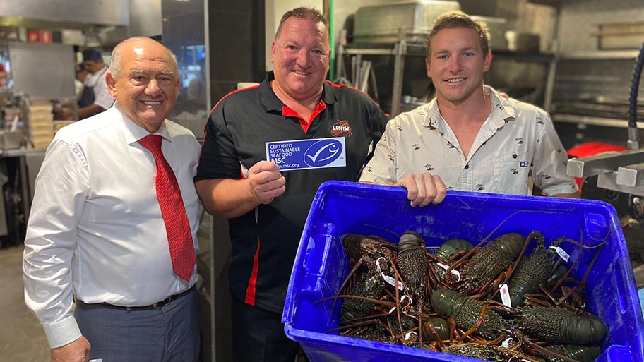 Three men standing, one holding MSC logo and one holding crate of rock lobsters   