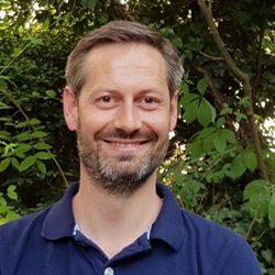 Head and shoulders shot of man in blue polo shirt in front of tree