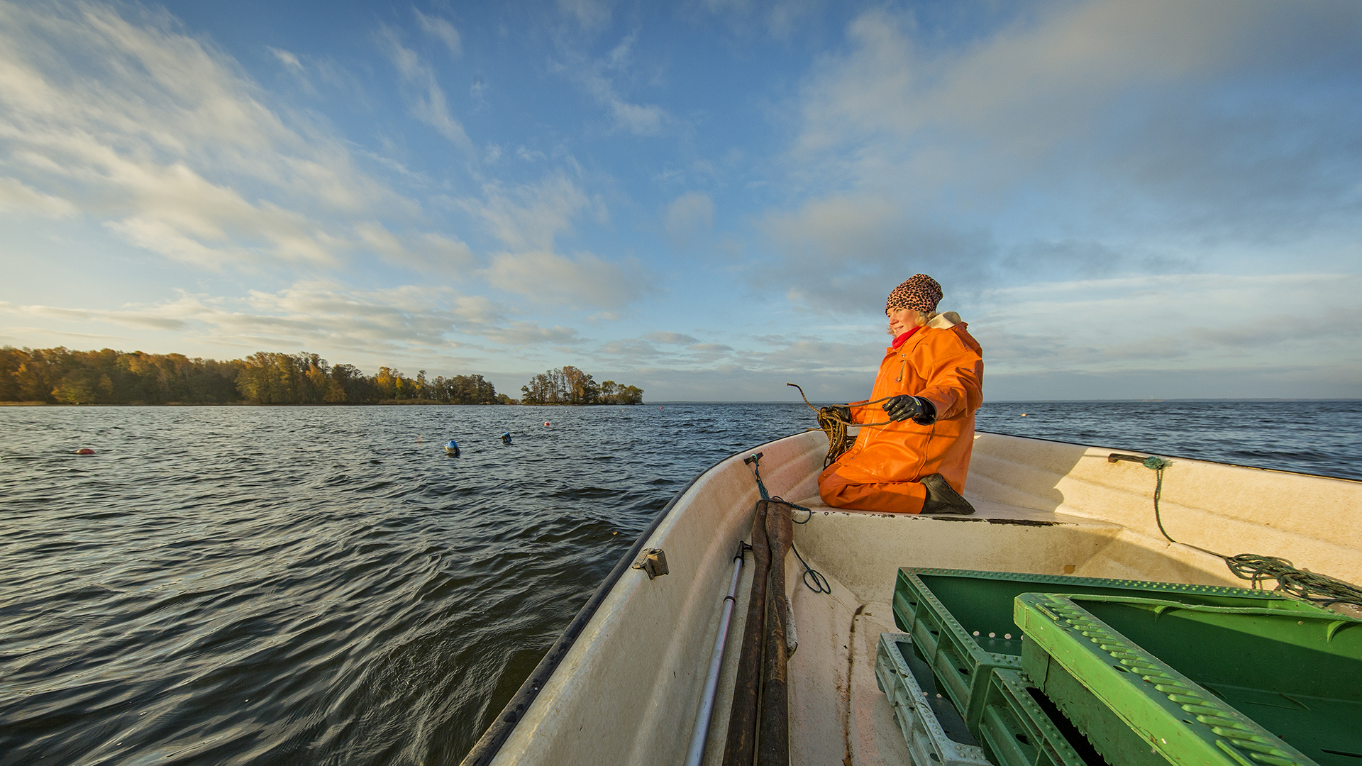 Woman fisher sitting on a boat in a lake. She is holding fishing line and there are trees in the background.