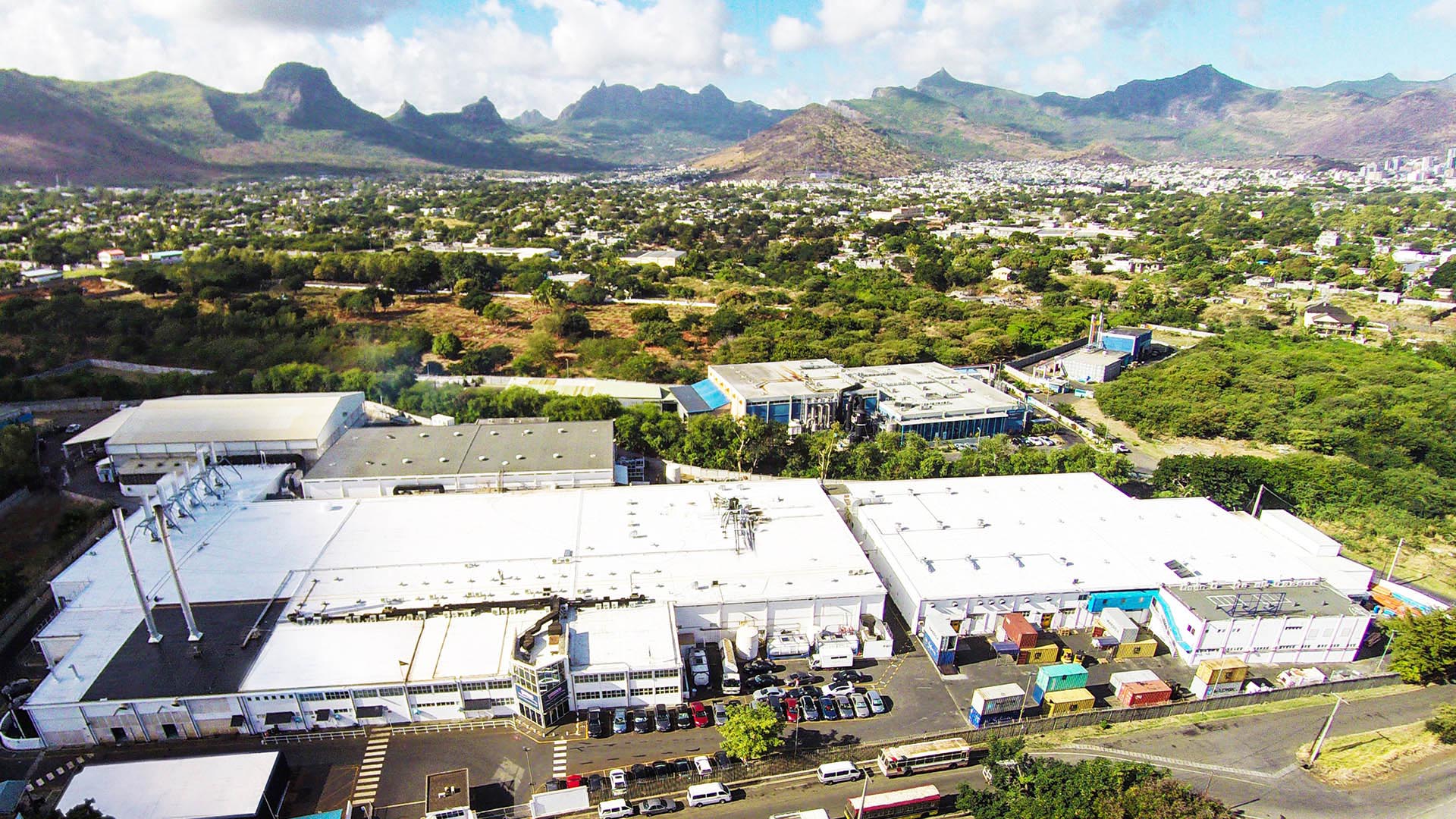 Wide view of white factory building, surrounded by trees, fields and small buildings with mountains and blue sky in the distance