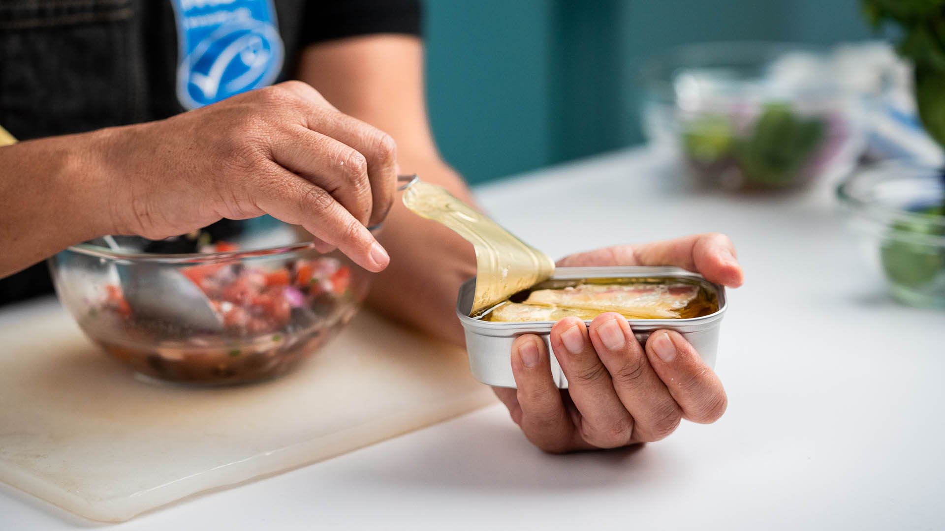 Woman's hands opening tin of sardines on counter, with bowl and MSC logo apron