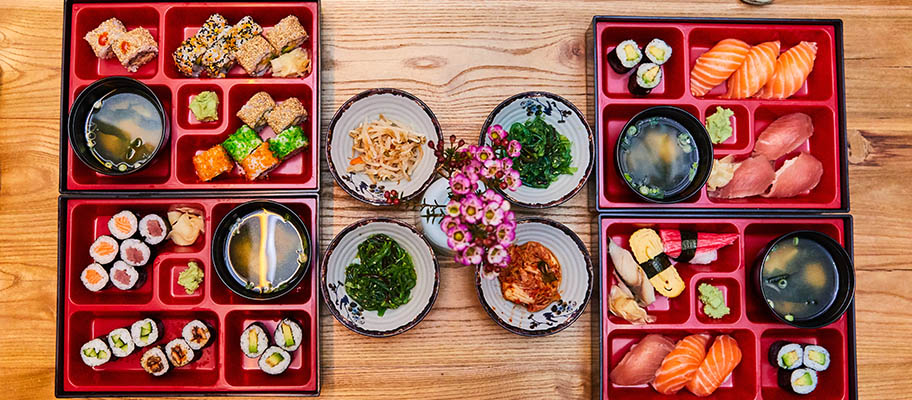 Two Japanese bento boxes from above with sashimi, nigiri, miso soup and plates of tsukemono pickles between