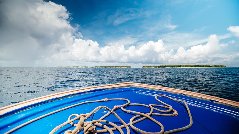 Bow of small blue fishing boat with ropes facing islands on horizon with blue sky and clouds