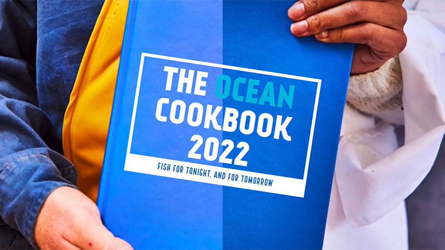 Blue book with text, THE OCEAN COOKBOOK 2022, held at corners by two different hands - one in chef's apron, one in fisher's overalls