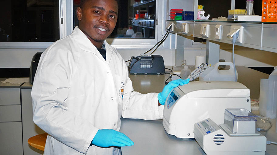 Young man in white labcoat and blue surgical gloves standing at counter with DNA extraction machinery