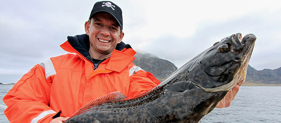 Man in baseball cap and orange waterproof jacket holding large halibut fish