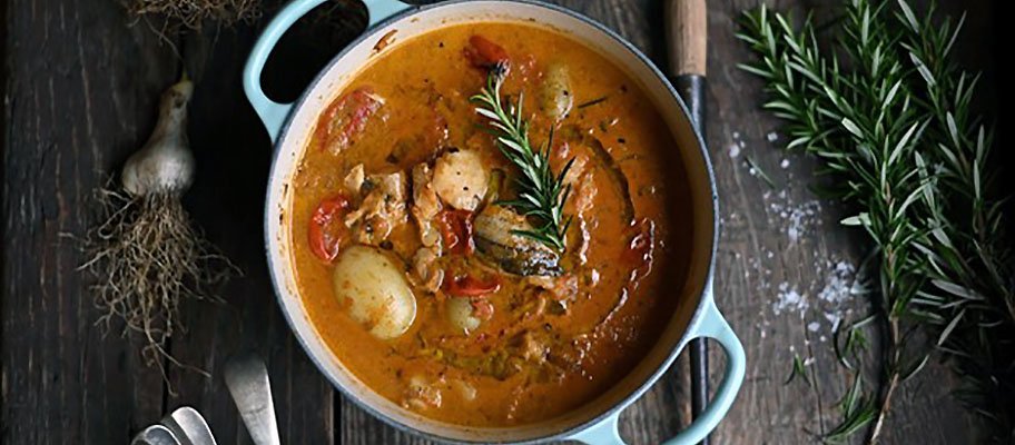 Pot of stew on wooden table seen from above. with sprigs of fresh herbs