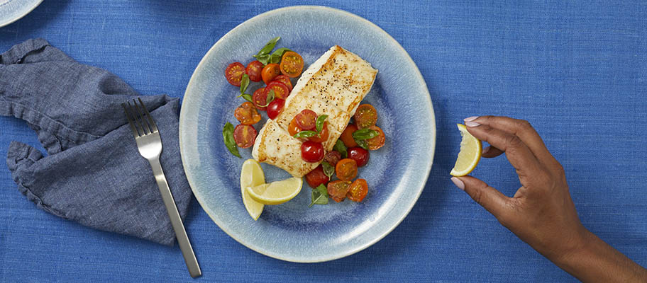 Plate of halibut with tomatoes and lemon slices on blue tablecloth with hand squeezing lemon