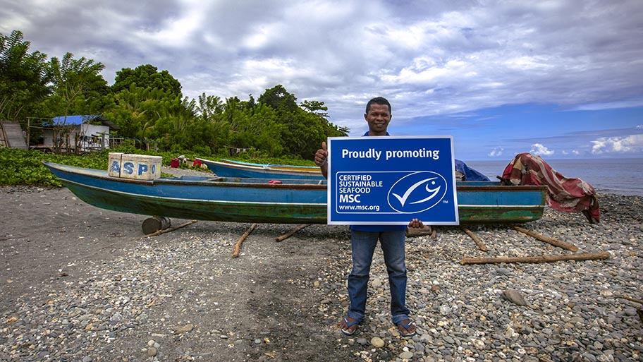 Man standing on shore in front of small boats, holding large sign with MSC logo