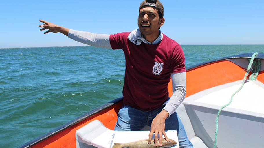 Man on small boat holding fish down and pointing out to sea