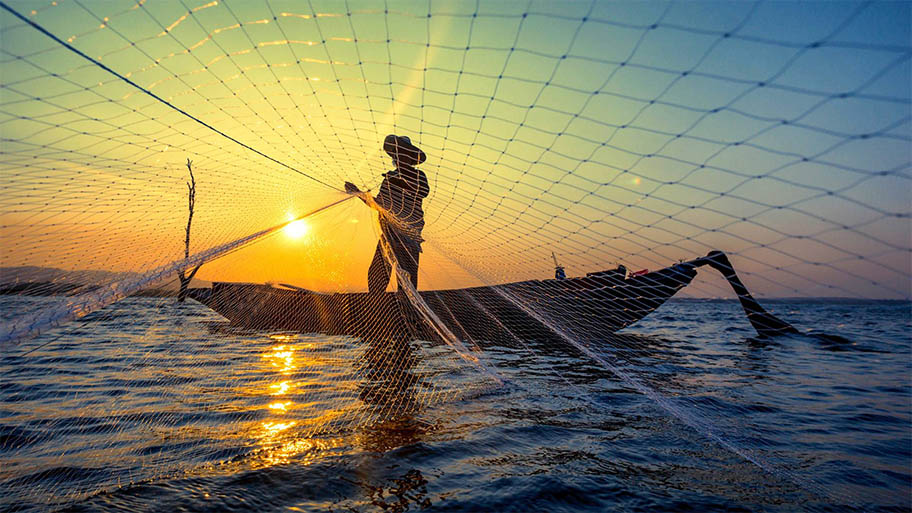 Fisherman in small boat throwing a net with the sunset behind him