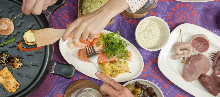 Raclette grill from above with hands reaching for salmon and prawns on side dish