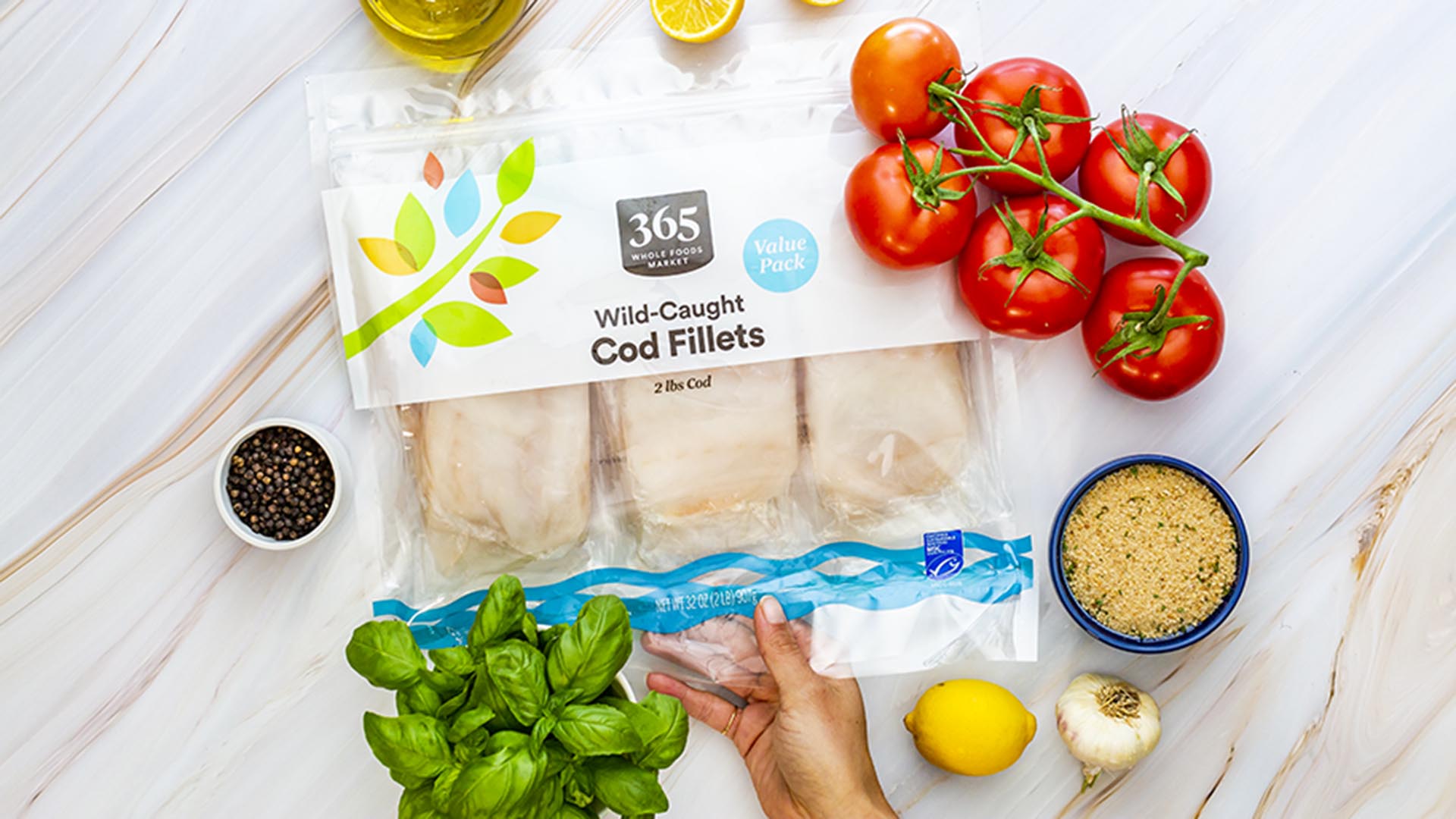 Hand reaching for cod fillets in packet on a table with tomatoes, garnish and dip