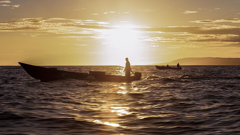 Silhouette of single fisher on small boat at sunset, with second boat on the horizon