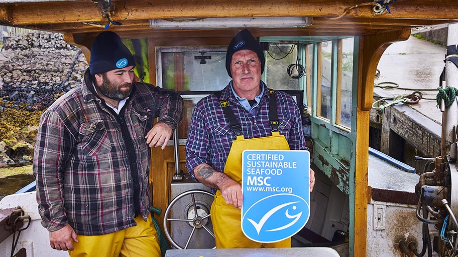 Two men in overalls on deck of small fishing boat holding MSC logo