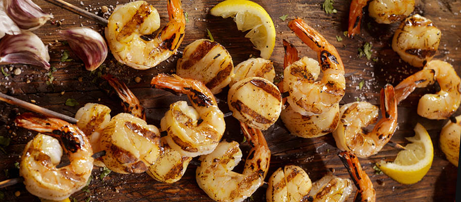 Grilled scallops and shrimp on wooden board with wooden skewers, chunks of lemon and garlic cloves