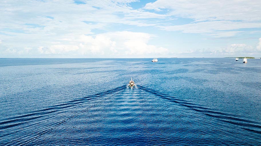 Small boat moving away from camera on blue water with blue sky and clouds, more small boats on horizon