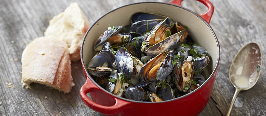 Steamed mussels in red pan with spoon and bread on wooden surface