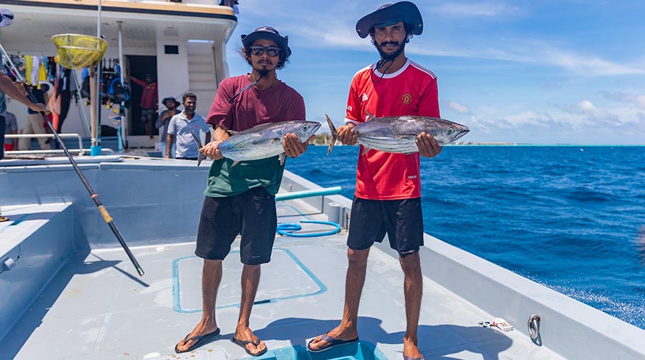 Two men standing on deck of boat, each holding a tuna fish. Blue sky and sea behind.