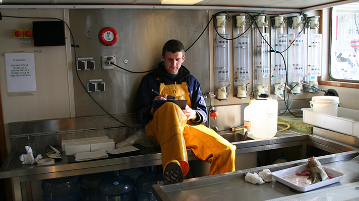Scientist sitting in a laboratory on  a research vessel making notes on a clipboard. 