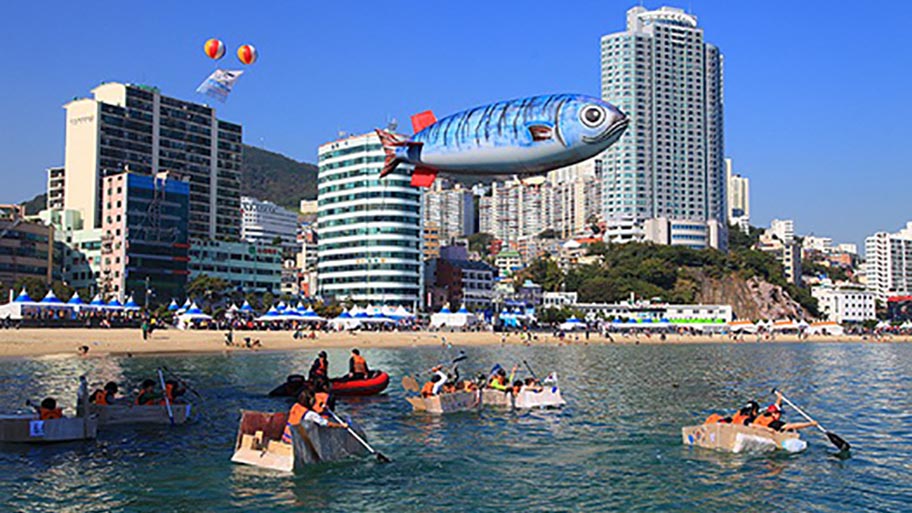 People in small rowing boats with inflatable mackerel above and buildings behind