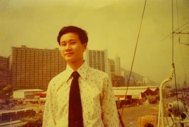 Old colour photograph of man in white shirt and dark tie with fishing wharf behind