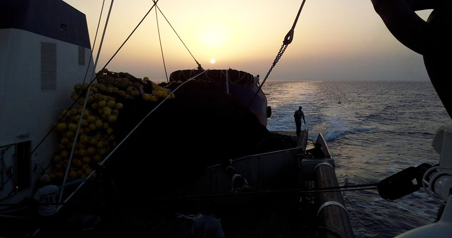 Silhouette of fishing gear and man on deck of large ship at sea facing setting sun