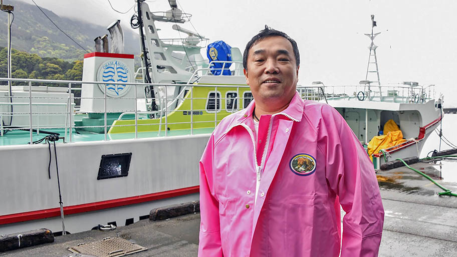 Man in pink waterproof jacket smiling in front of fishing vessel
