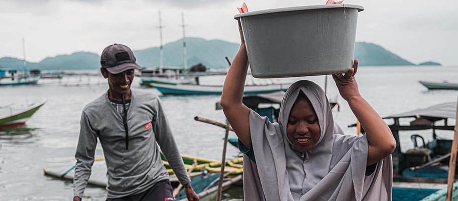 Man and woman stepping off small fishing boat, woman carrying bucket of fish on her head