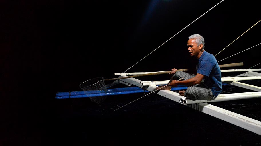 Fisher sitting on frame of boat with handline and net, dark background