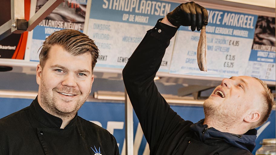 Two fishmongers in dark uniforms, one holding herring by its tail above his mouth