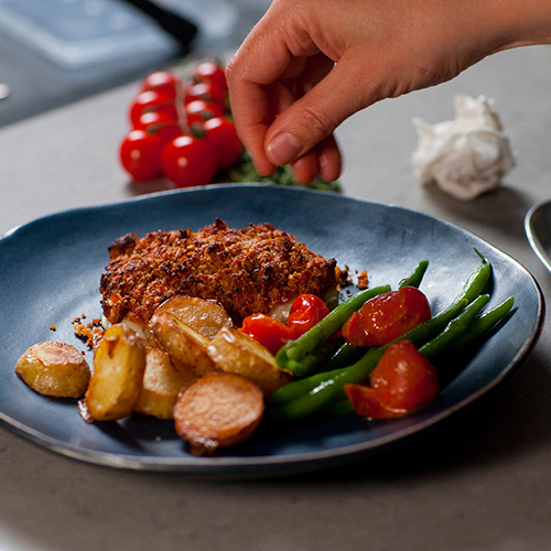 Person's hand sprinkling garnish on to a plated dish of crusted baked cod