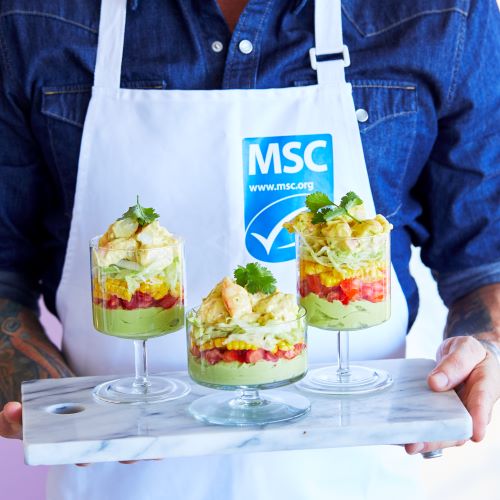 Three parfait glasses stacked with prawns lettuce corn tomato and avocado on a marble board held by a person wearing a blue denim shirt and a white apron with a blue label saying MSC www.msc.org