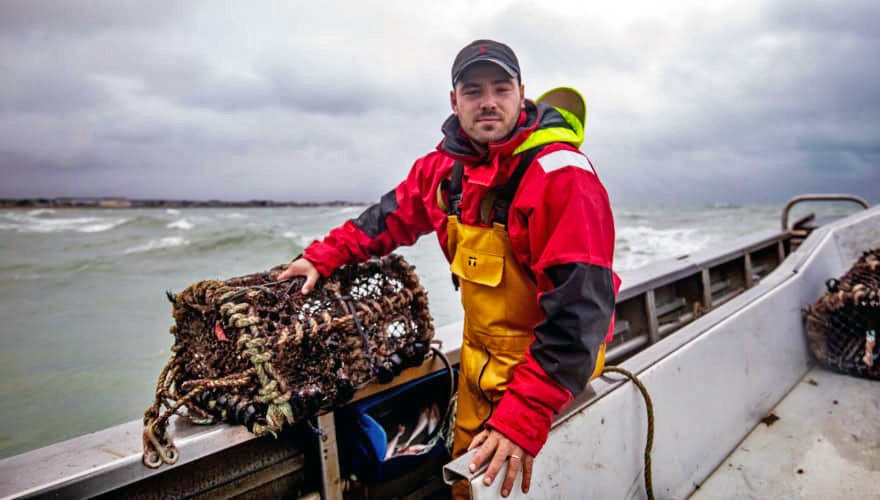 Fisher in red jacket in boat at sea, holding lobster trap on the edge of the boat