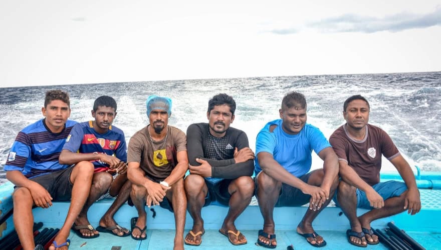 Six fishermen in shorts and flip-flops sitting on the back of a boat.