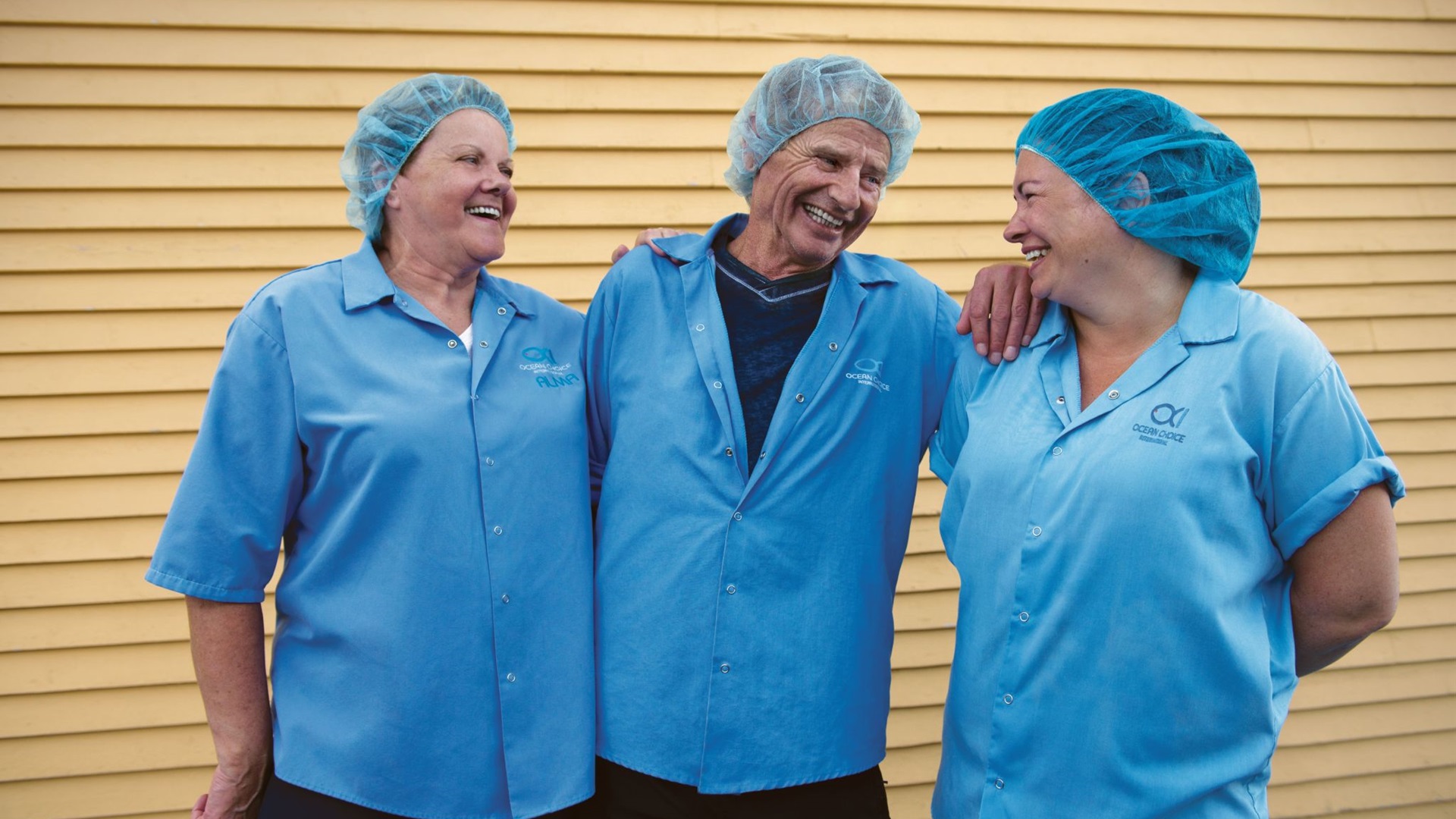 Three people smiling at each other wearing blue button down shirts and blue hair nets.