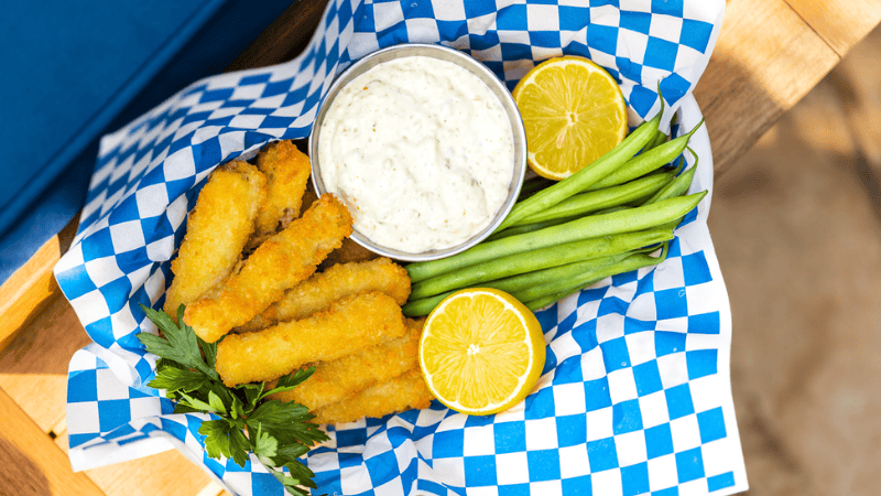 Based of fried fish sticks with a cup of tartar sauce and green beans in a basket with blue checkered paper