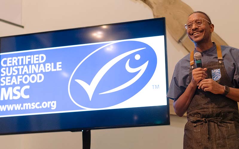 Chef Gregory Gourdet smiles while holding microphone in front of large screen