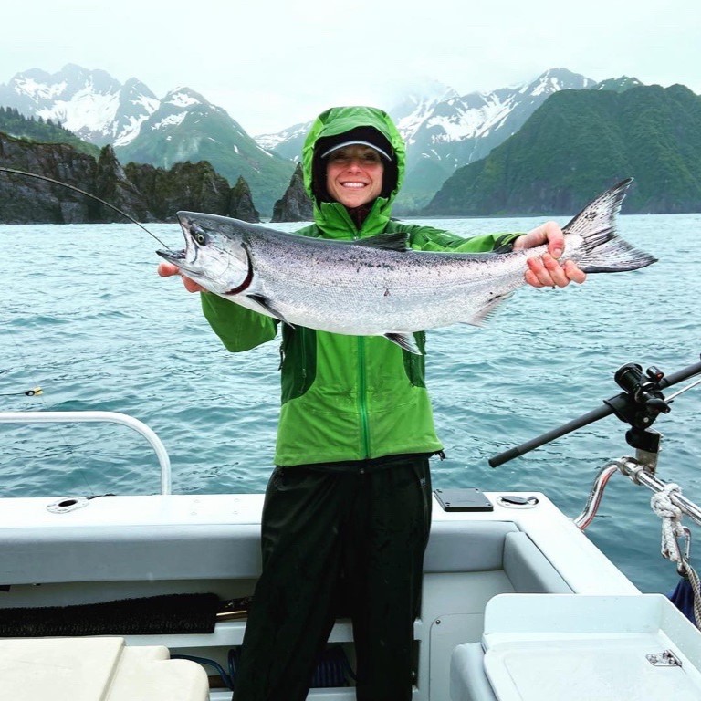 MSC staff member Laura McDearis on a boat holding a salmon she caught in front of snow capped mountains.