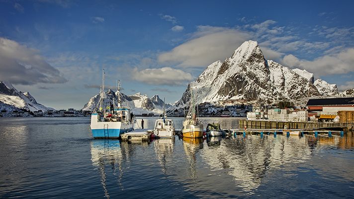 Fiskebåtar i förgrunden. Snöklädda berg och blå himmel i bakgrunden.