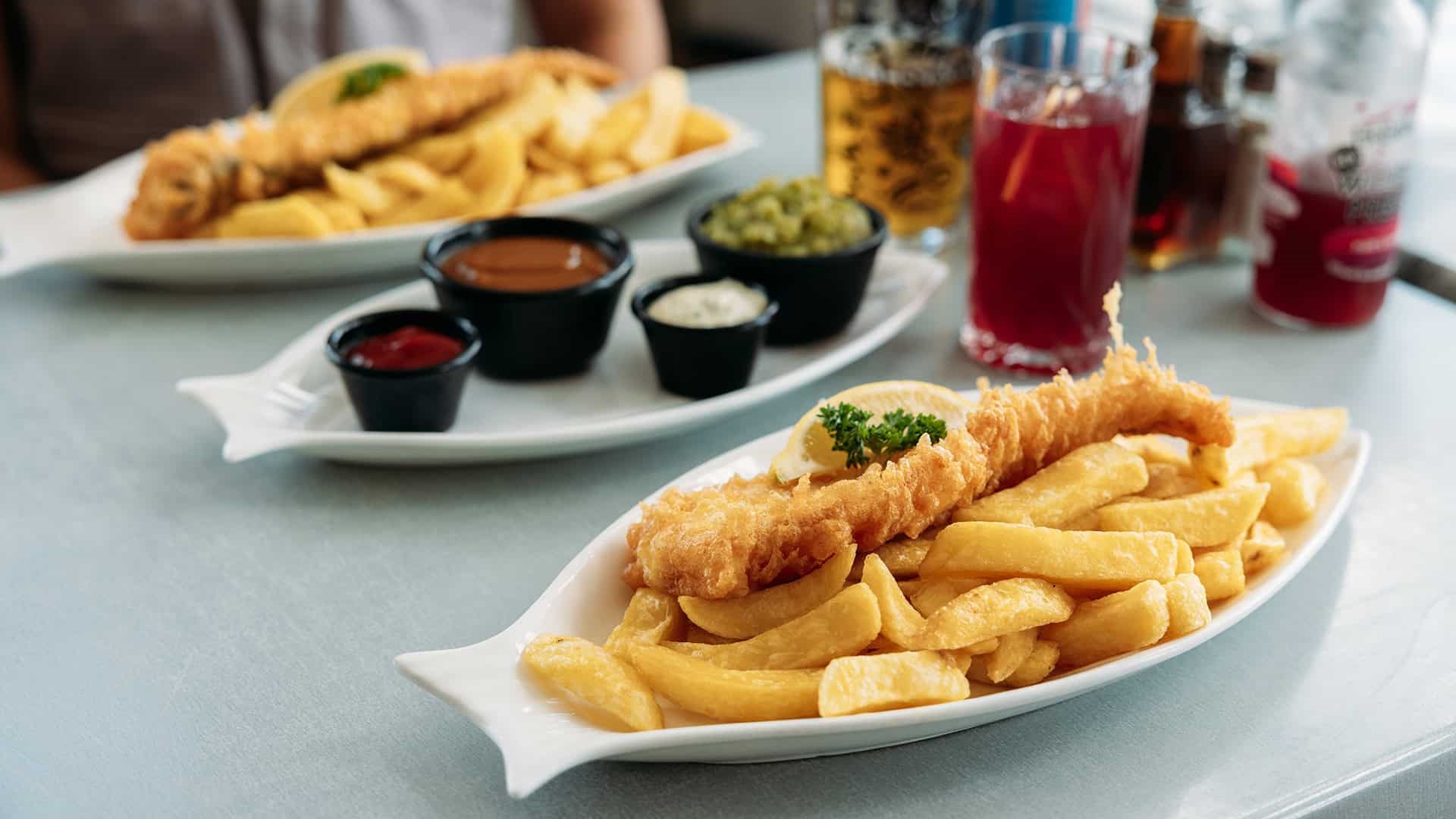 Portion of two fish and chips on plates with sauces and drinks